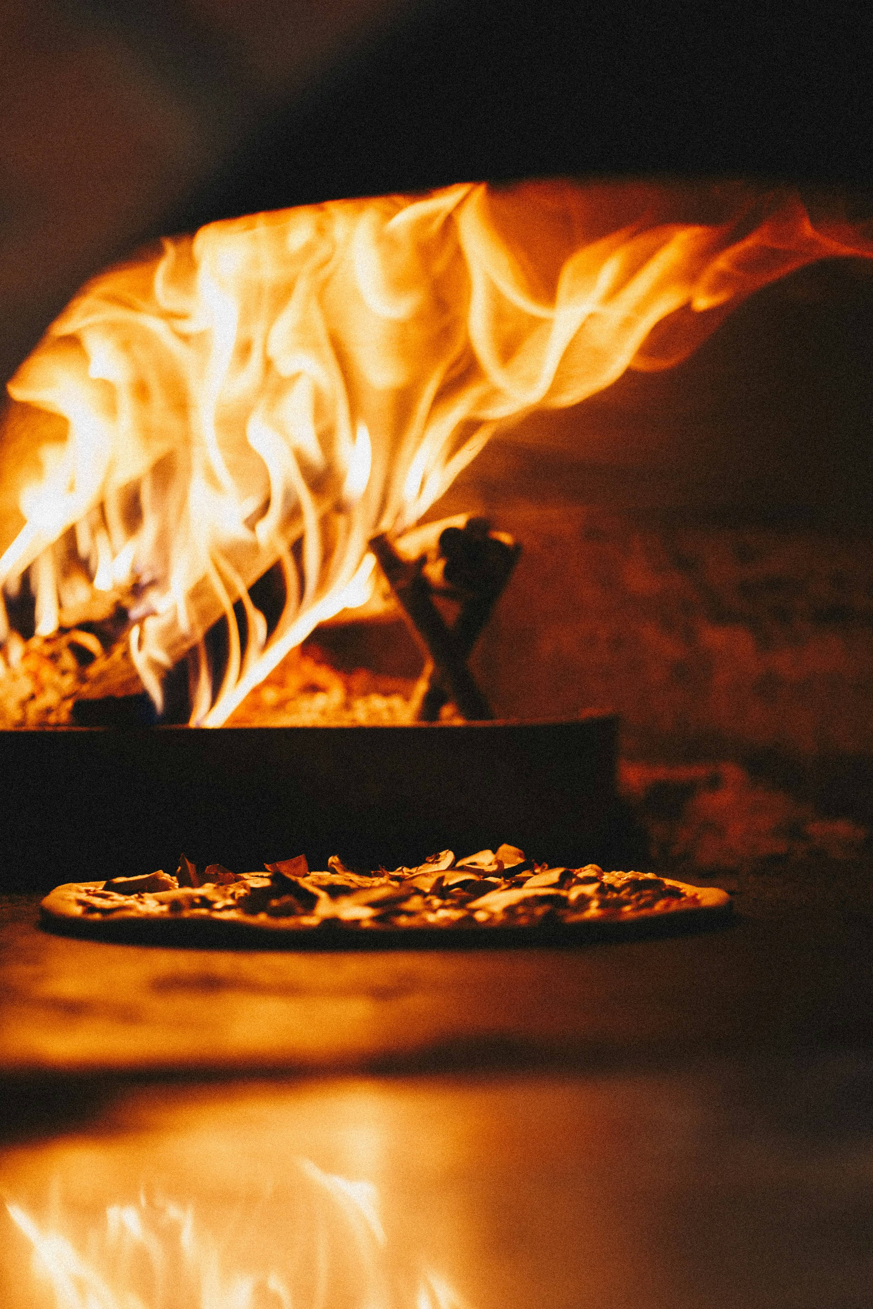 Fiamma alta nel forno a legna con una pizza in cottura in primo piano
