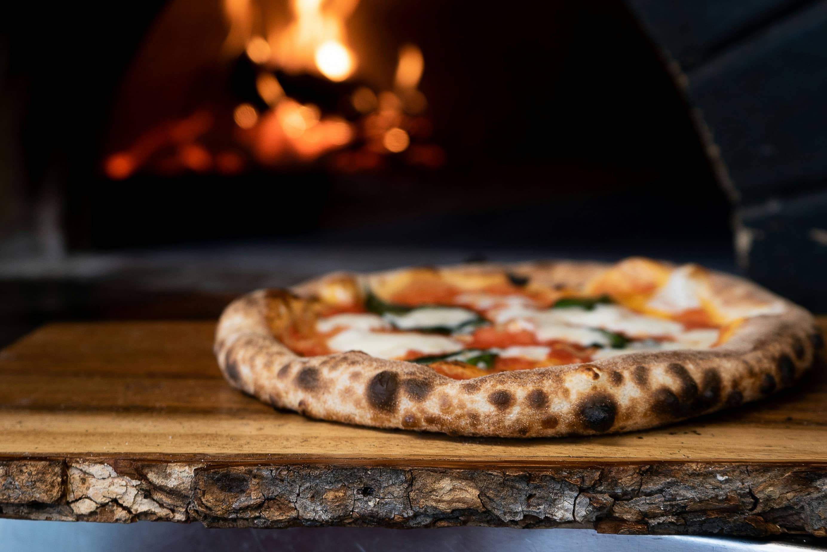 Pizza con pomodoro, fior di latte e basilico su tagliere di legno, forno acceso alle spalle